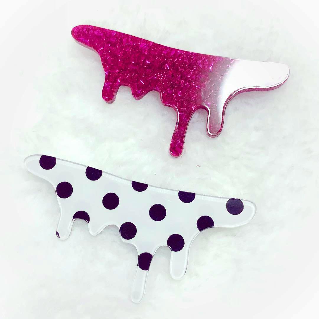 Two drippy-shaped plastic brooches shown on a white background, one pink glitter and one white with black polka dots.
