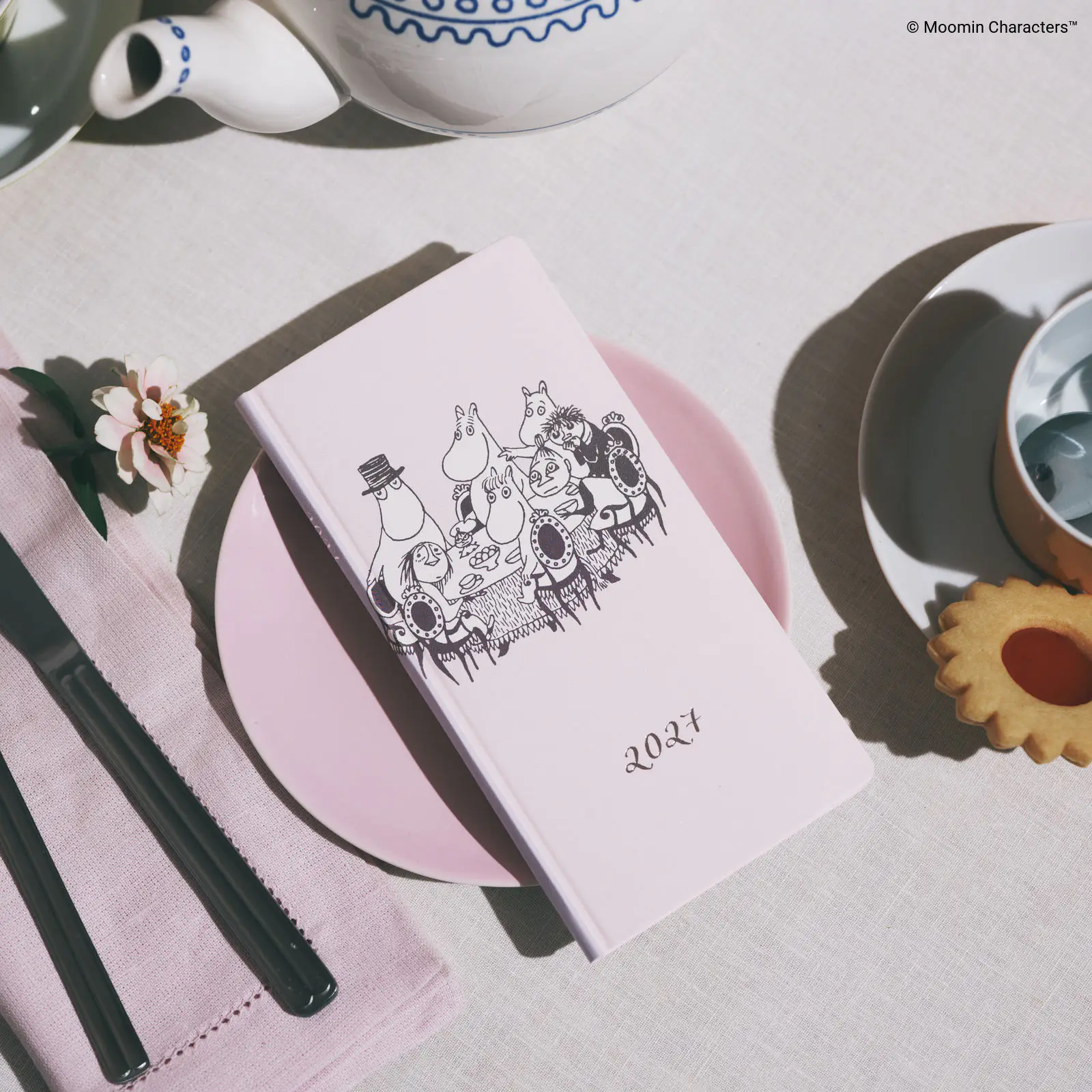 White Moomin-themed weekly notebook with a table illustration placed on a pink plate beside a teapot, cutlery, and a cookie.