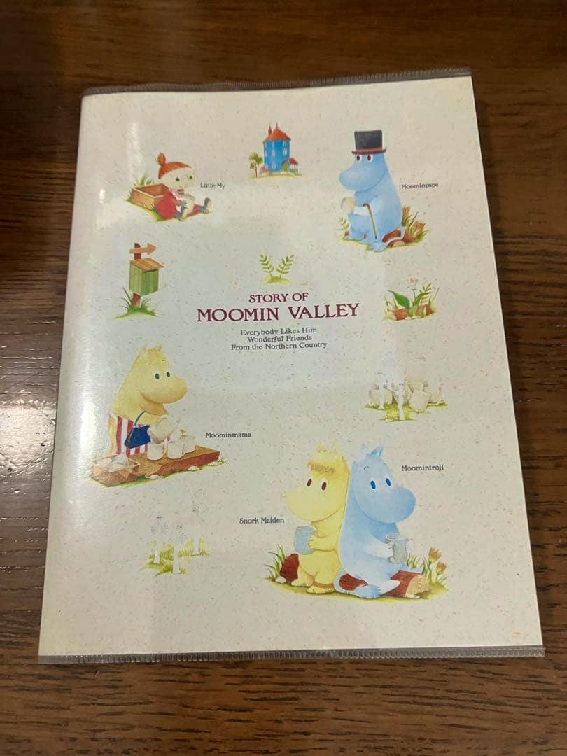 Front cover of the Moomin mini notebook titled "STORY OF MOOMIN VALLEY" with illustrated Moomin characters on a wooden surface.