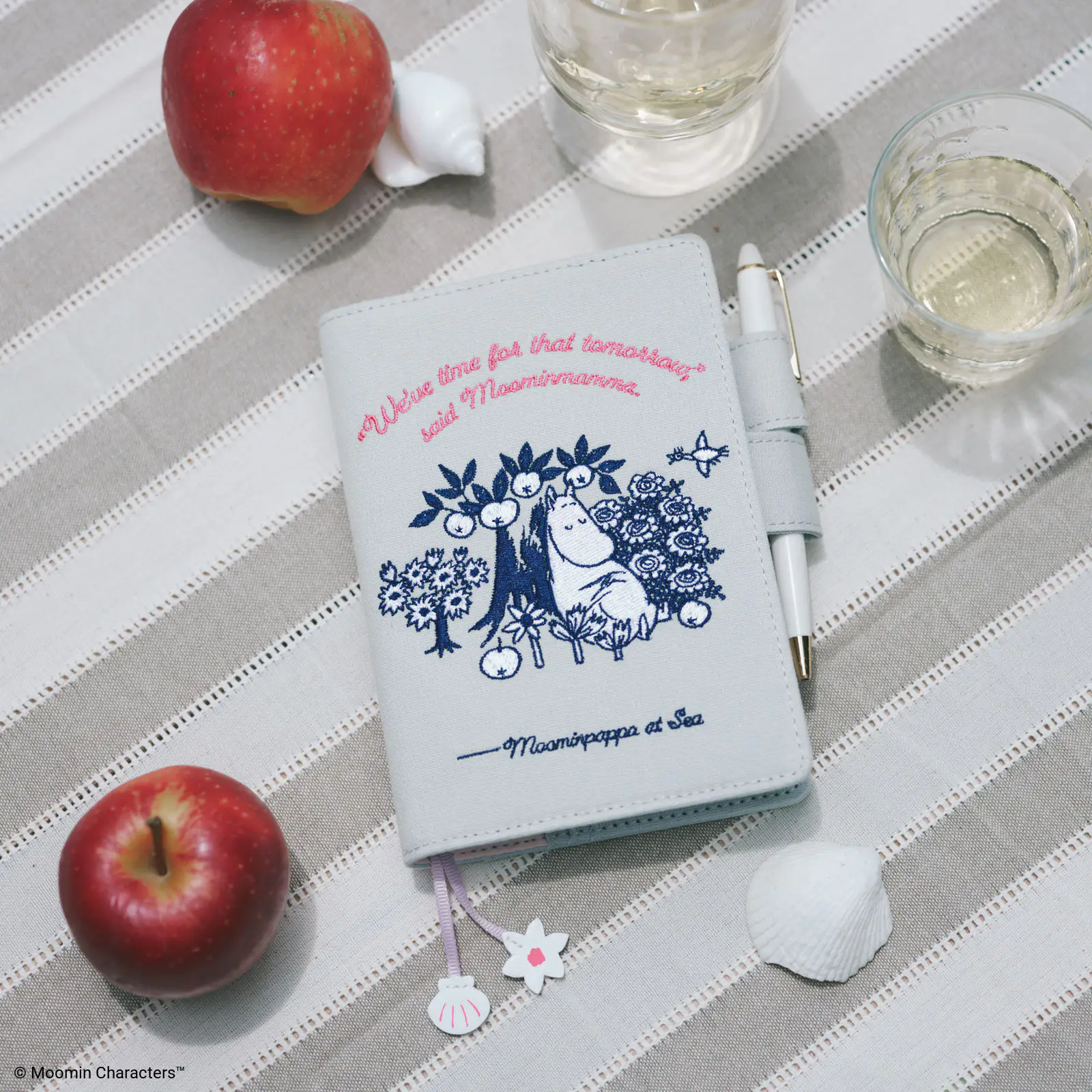 Pale mint A6 diary with Moomin "My Yard" cover design, pen attached, displayed on a striped cloth with apples and glasses.