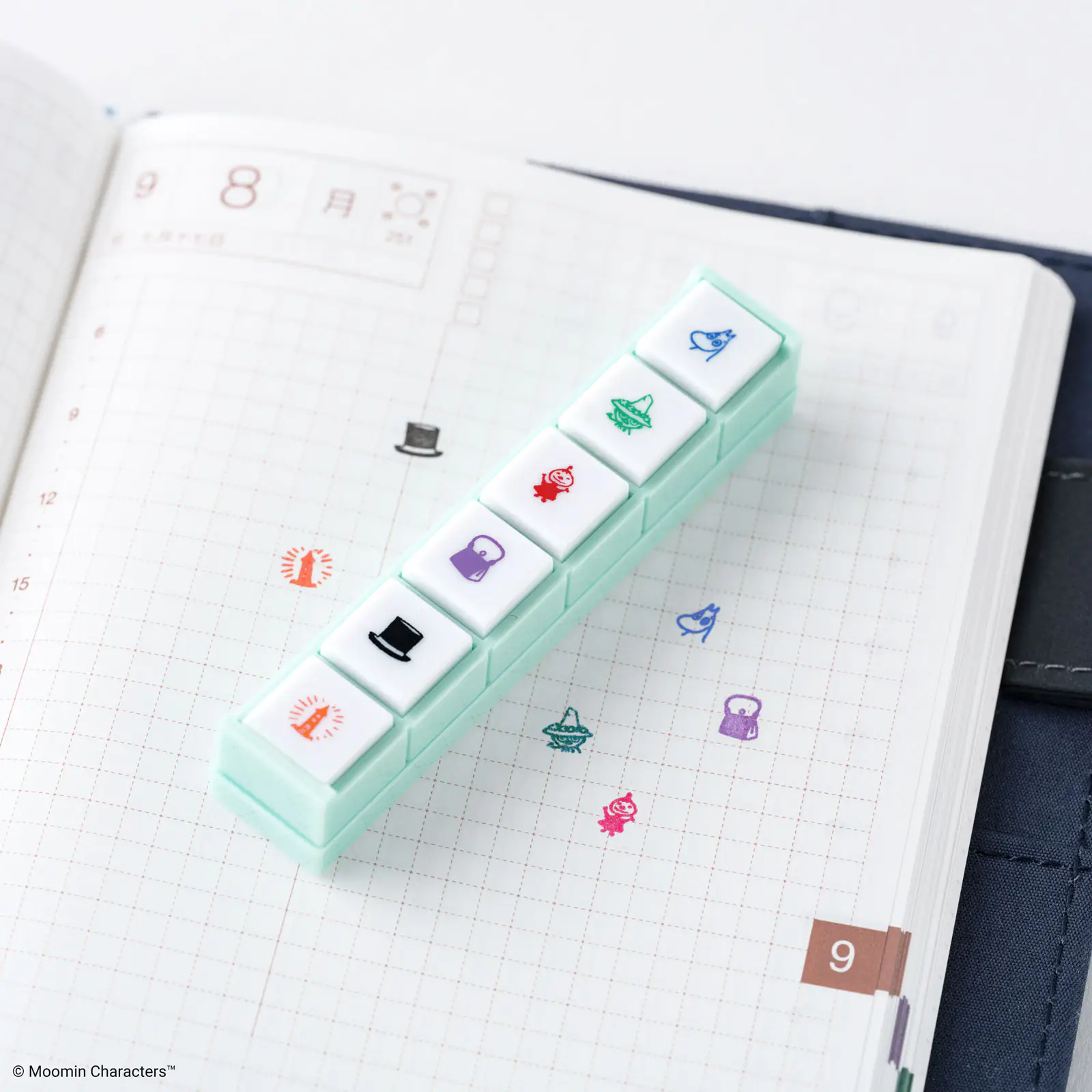 A mint-green stack of six push-button stamps with Moomin icons placed on a grid notebook showing colorful stamped icons.
