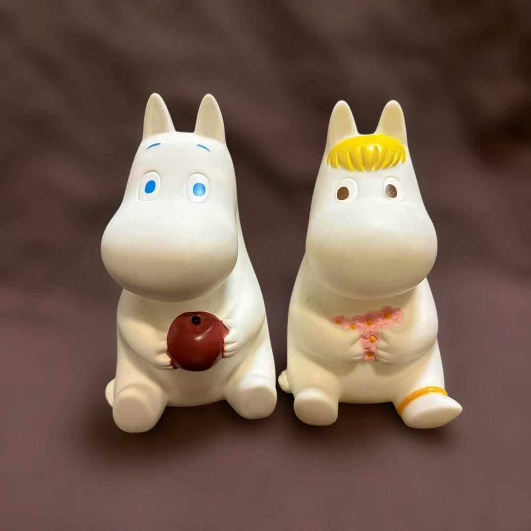 Front view of two white Moomin soft-vinyl piggy bank figures sitting side by side; the left holds a red apple and the right holds pink flowers.
