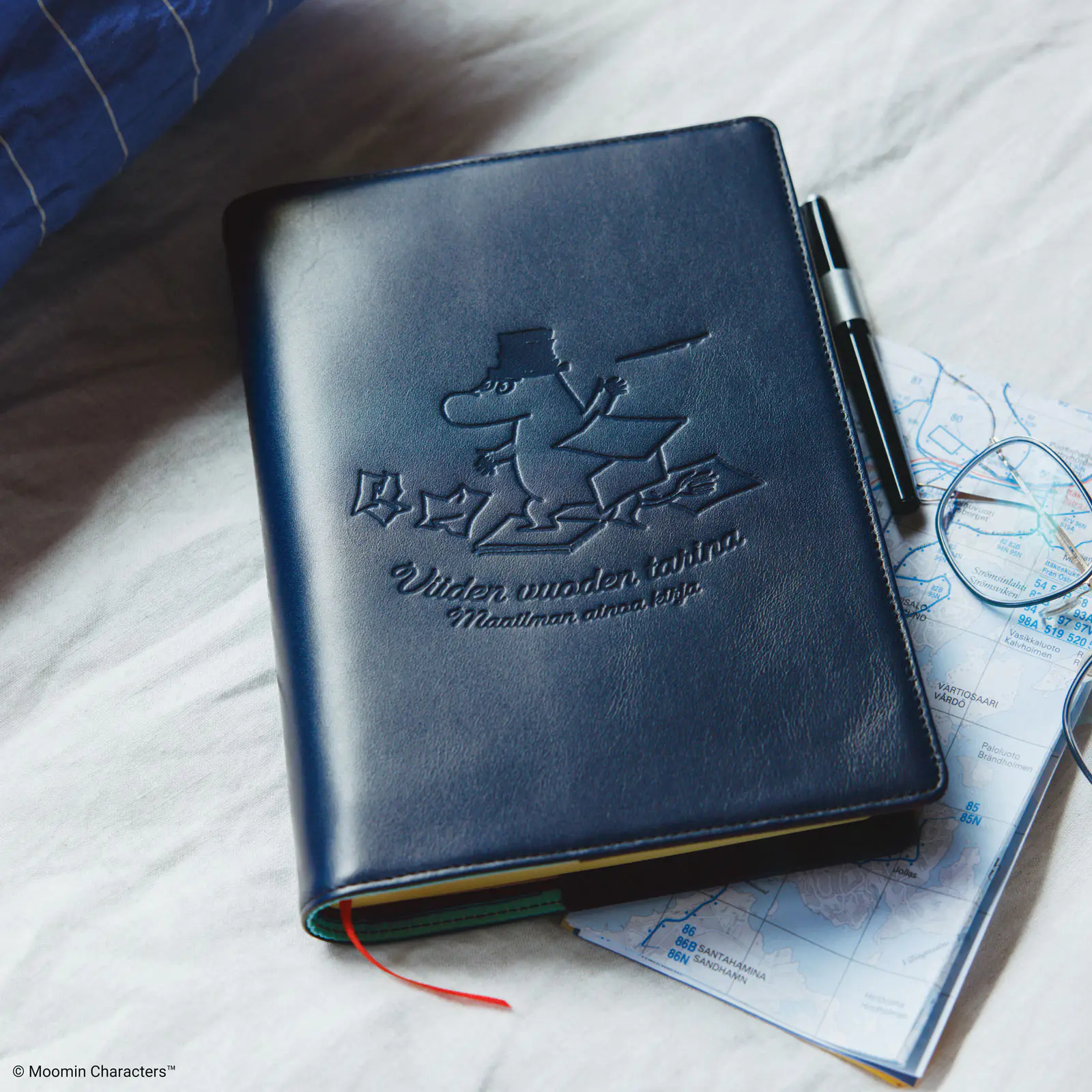 Dark blue cow leather book cover for a large 5-year techo with an embossed Moomin character, shown on a surface with a pen and a map nearby.
