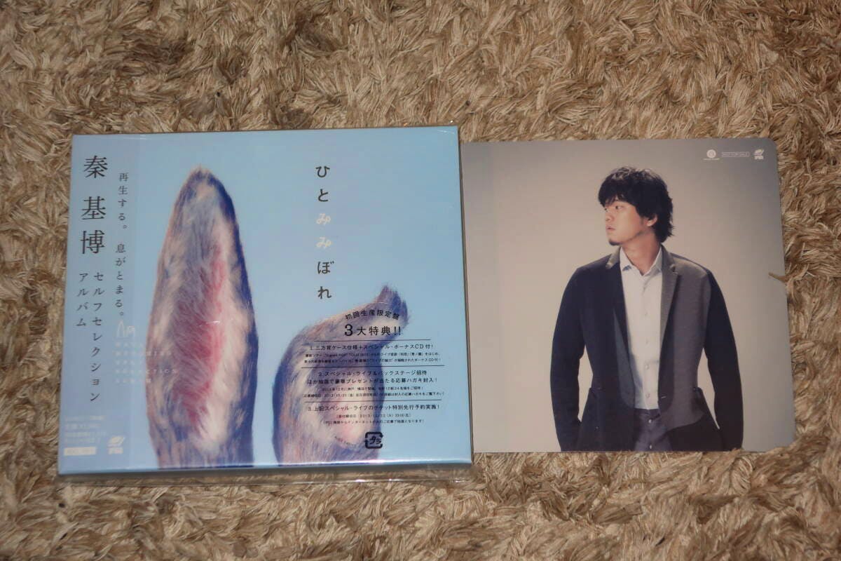 Sealed CD package of 秦基博 "ひとみみぼれ" with blue cover art and an included artist photo jacket placed on a carpet.