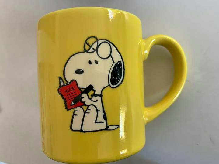 Yellow ceramic mug showing a Snoopy cartoon holding a red notebook, handle visible on the right.
