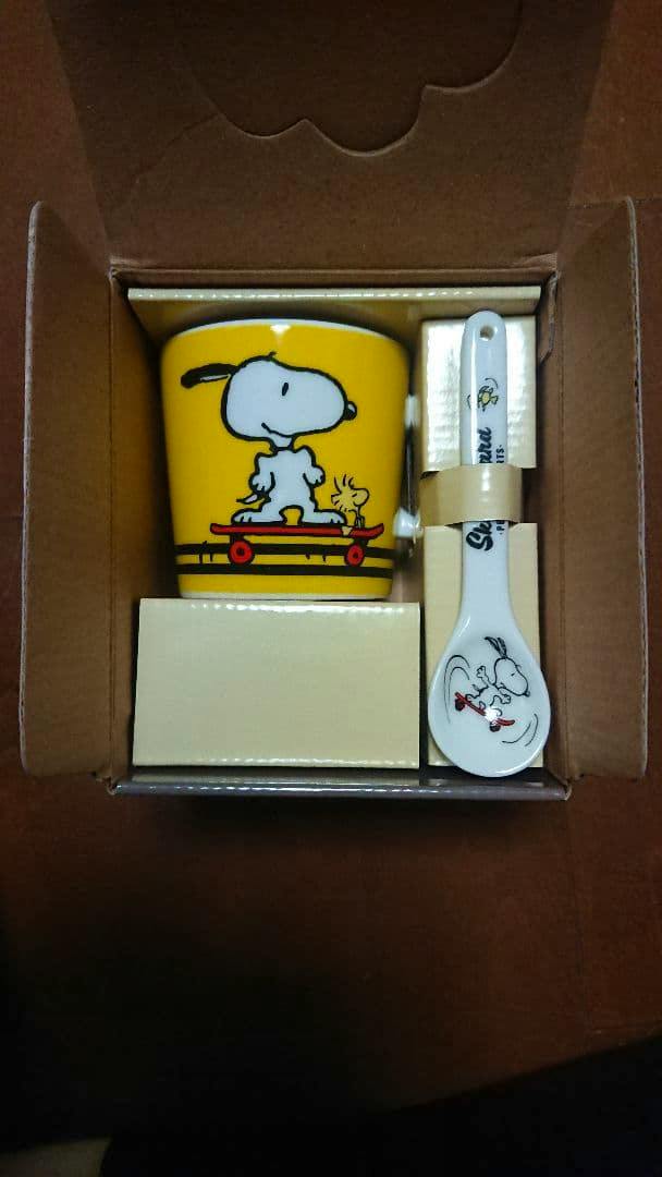 A yellow mug featuring Snoopy with a matching spoon shown inside a cardboard box.