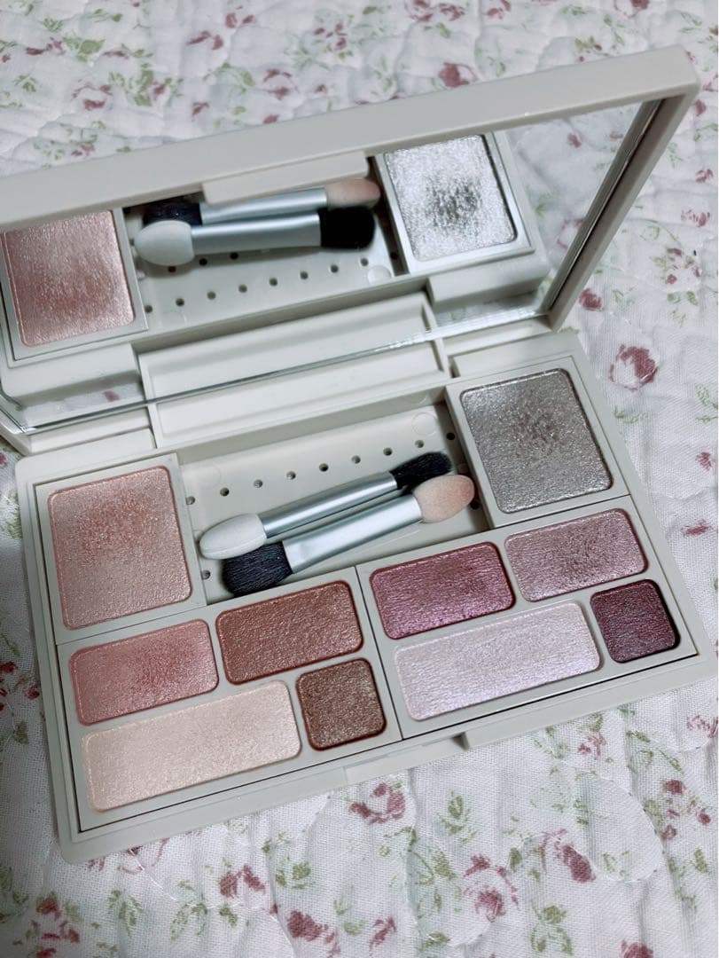 Open MUJI eye color palette showing multiple eyeshadow pans, mirror, and two sponge applicators on floral fabric.