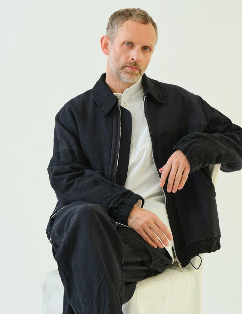 Male model seated wearing the men's wool multi-fabric patchwork zip-up short jacket over a white shirt.