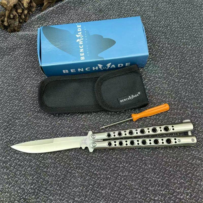 An open stainless steel butterfly pocket knife with a Benchmade box, black pouch, and small screwdriver on a dark textured surface.