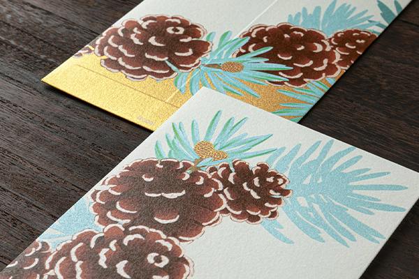 Close-up of multipurpose envelopes showing a pinecone wreath print and a gold-foiled edge.