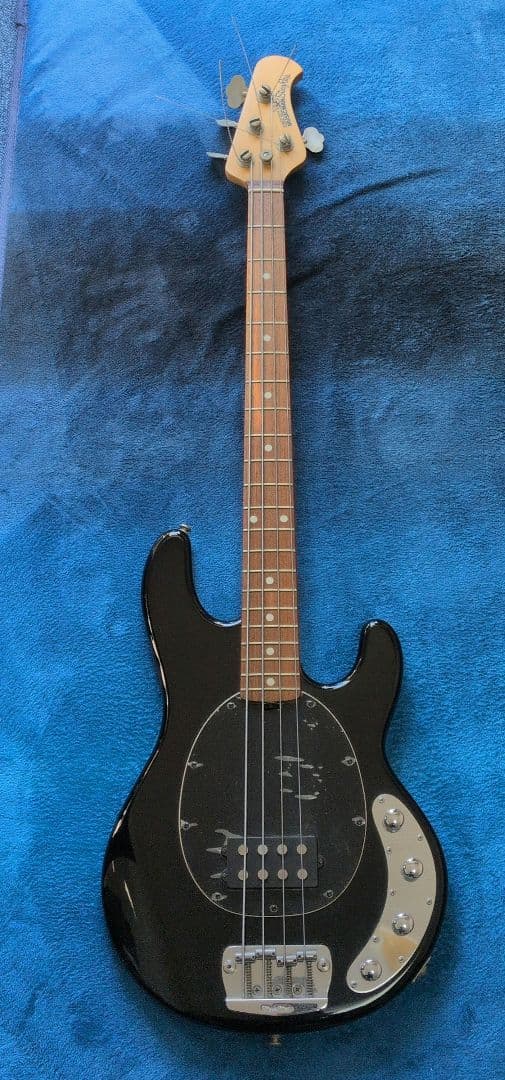Full-body photo of the black Musicman StingRay electric guitar on a blue fabric background, showing the body, neck, and bridge.
