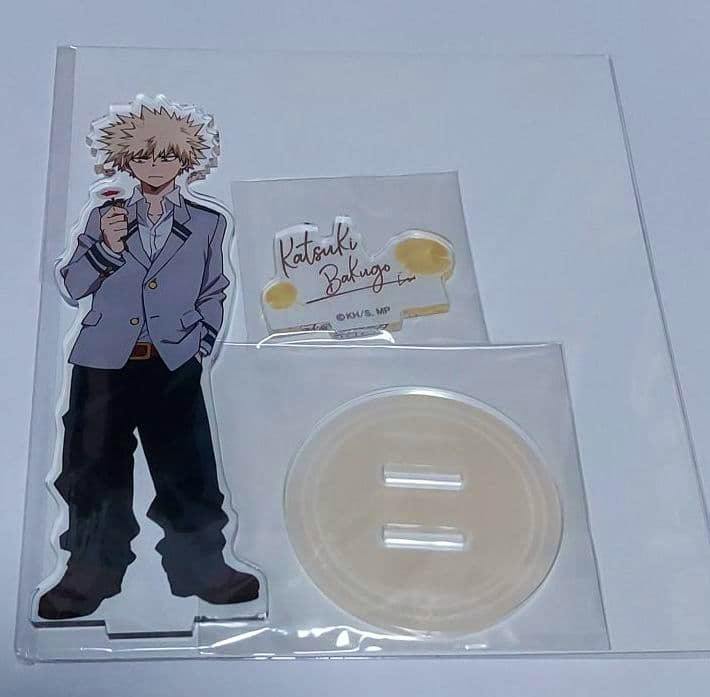 Packaged acrylic stand set showing a Katsuki Bakugo character standee, a small nameplate, and a round base in plastic packaging on a white background.