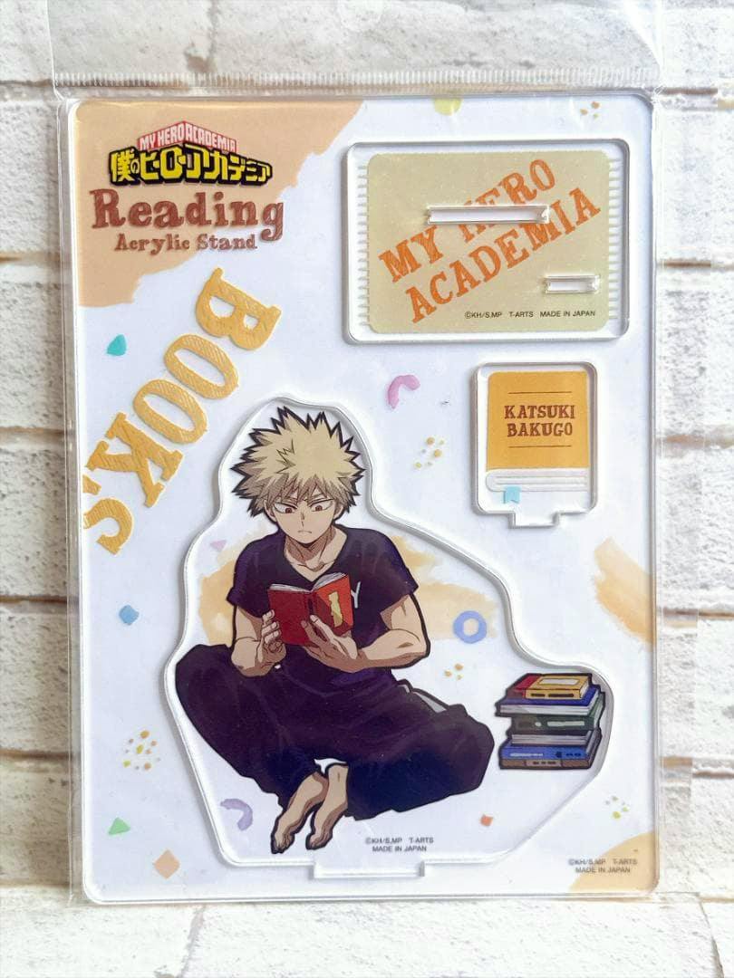 Packaged acrylic full-body stand showing Katsuki Bakugo reading a book with the stand base and accessory pieces visible in the blister pack.