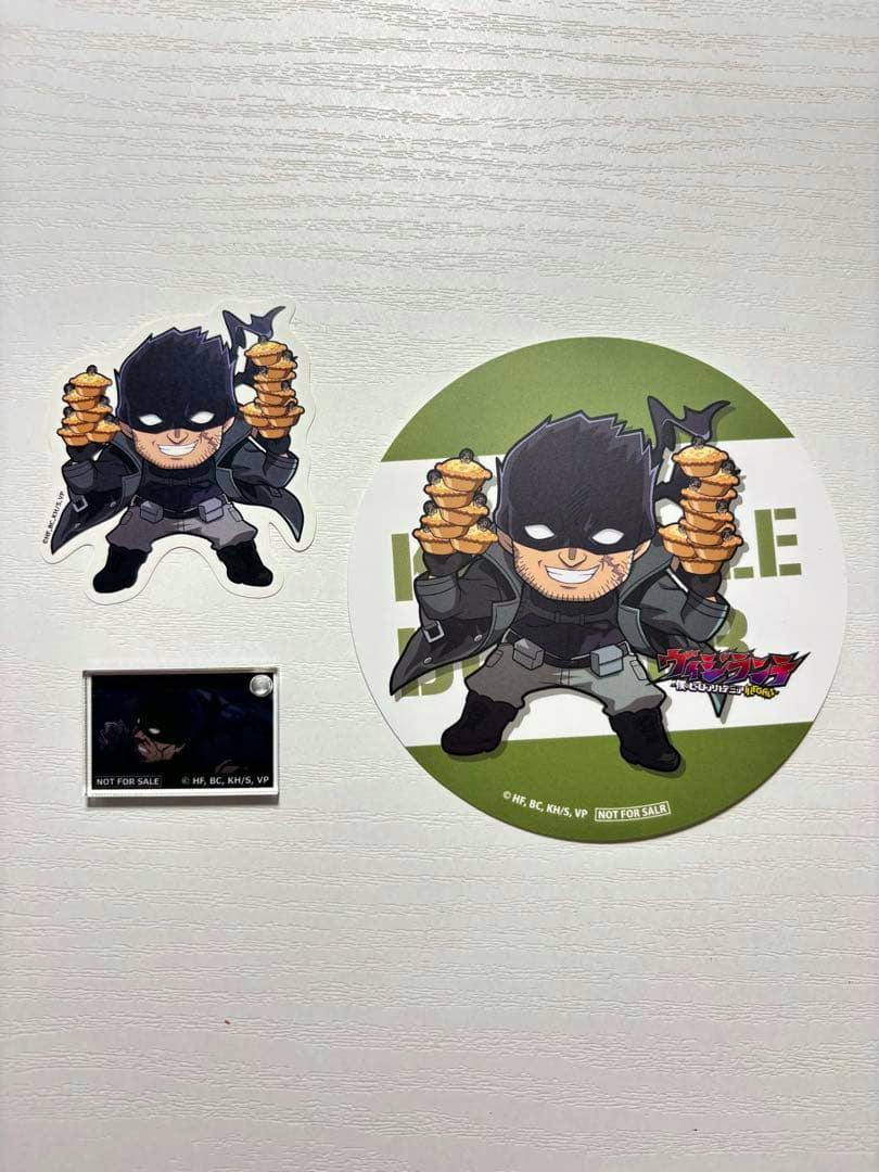 Photo of three Vigilante-themed items: a character sticker, a small acrylic keychain, and a round coaster displayed on a white background.