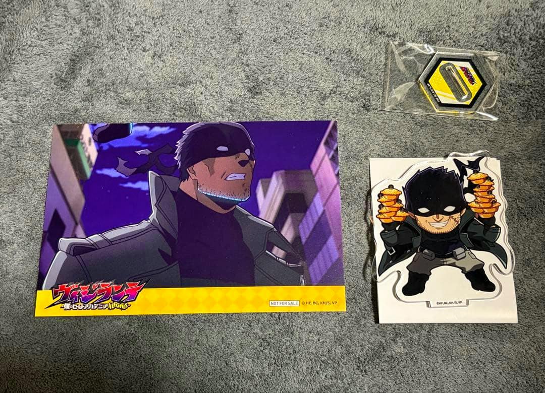 Flat lay of Vigilante PABLO mini merchandise on gray fabric: a large character print, a chibi PABLO acrylic with knuckle-duster props, and a small hexagonal packaged item.
