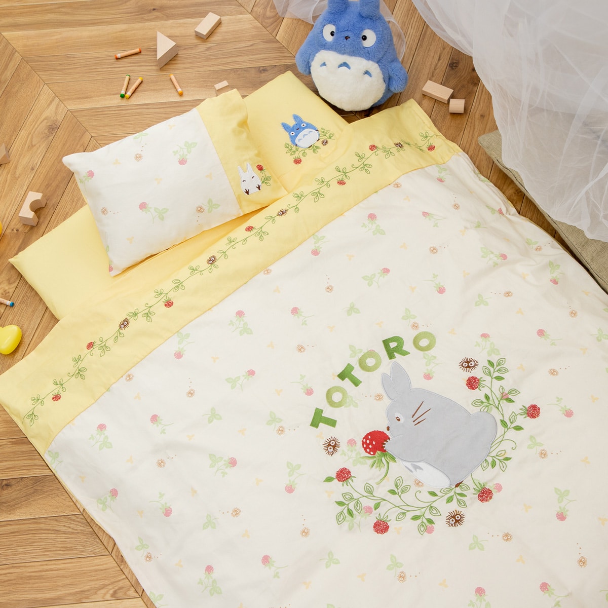 A baby futon set with Totoro and strawberry print on the comforter, with matching pillow and yellow sheets arranged on a wooden floor.