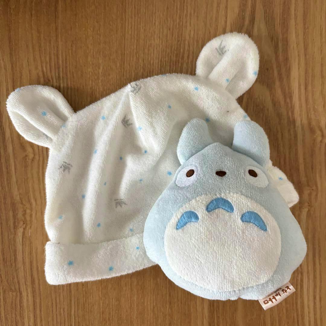 A white baby hat with small ears next to a light-blue Totoro rattle on a wooden surface.