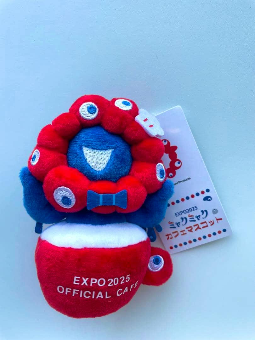 Front view of the red-and-blue plush keychain mascot showing embroidered white text "EXPO2025 OFFICIAL CAFE" and an attached product tag.