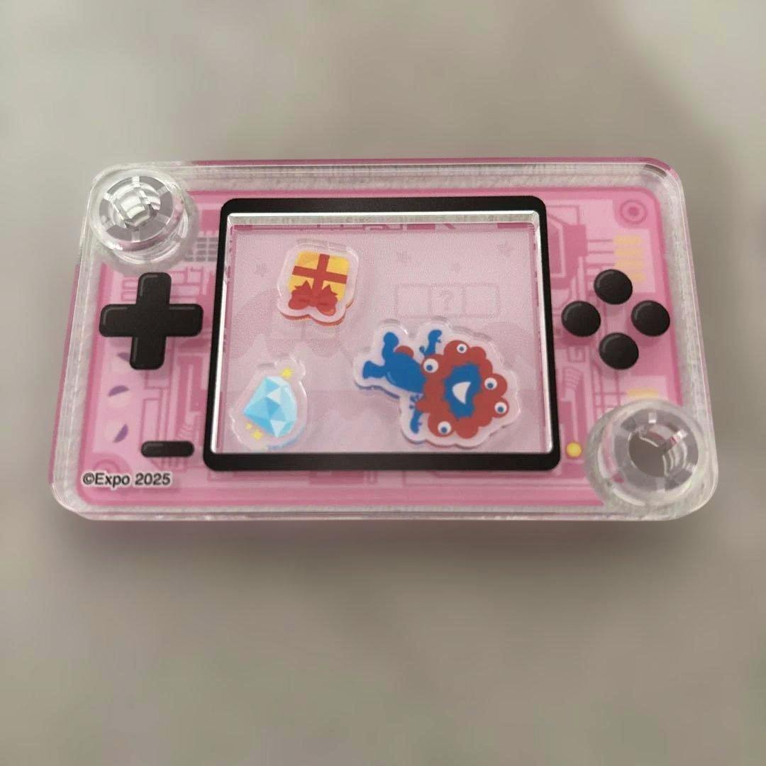 Front of a pink acrylic game machine–style keychain with a clear central window holding small stickers, a black D-pad at left and four black buttons at right.