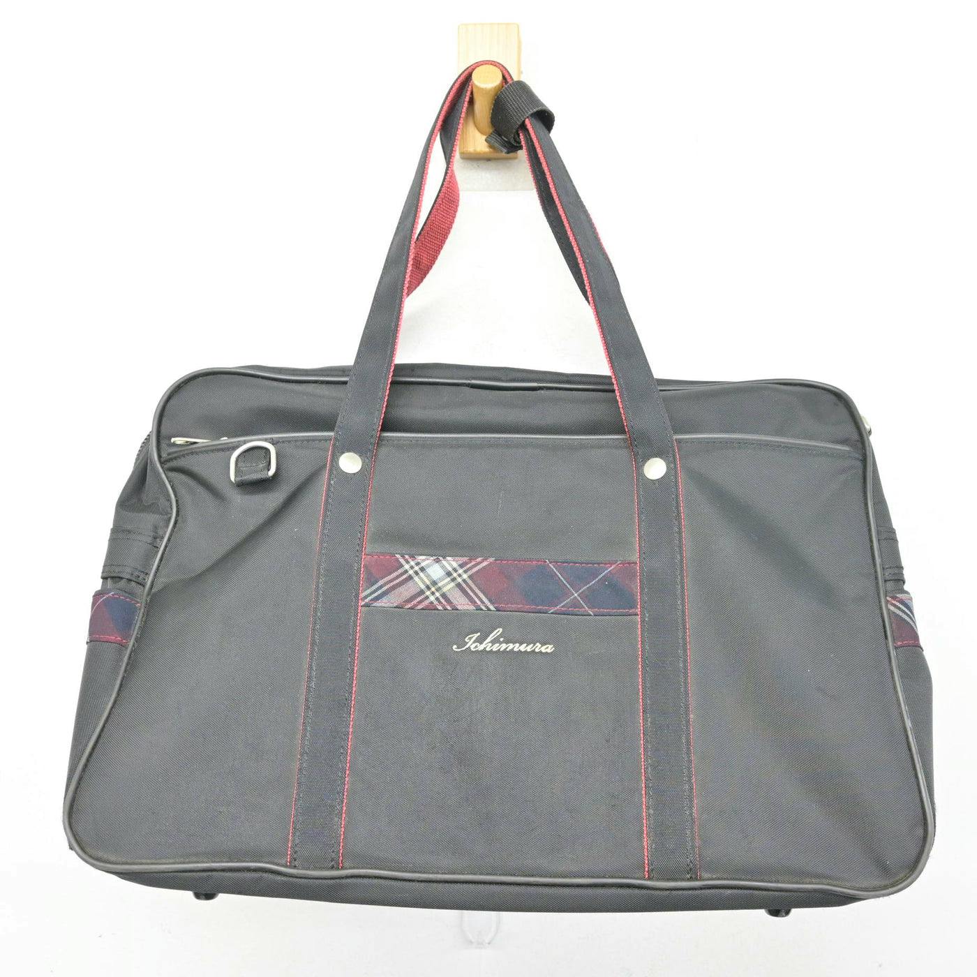 Black polyester bag with red-trimmed handles and a small plaid accent on the front, hanging from a wooden hook.