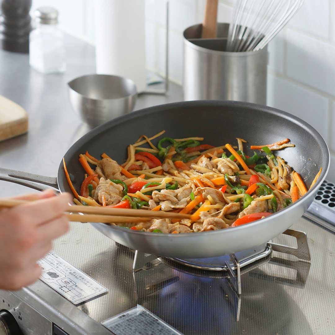 An iron frypan on a gas stove containing stir-fried vegetables and meat being stirred with chopsticks.