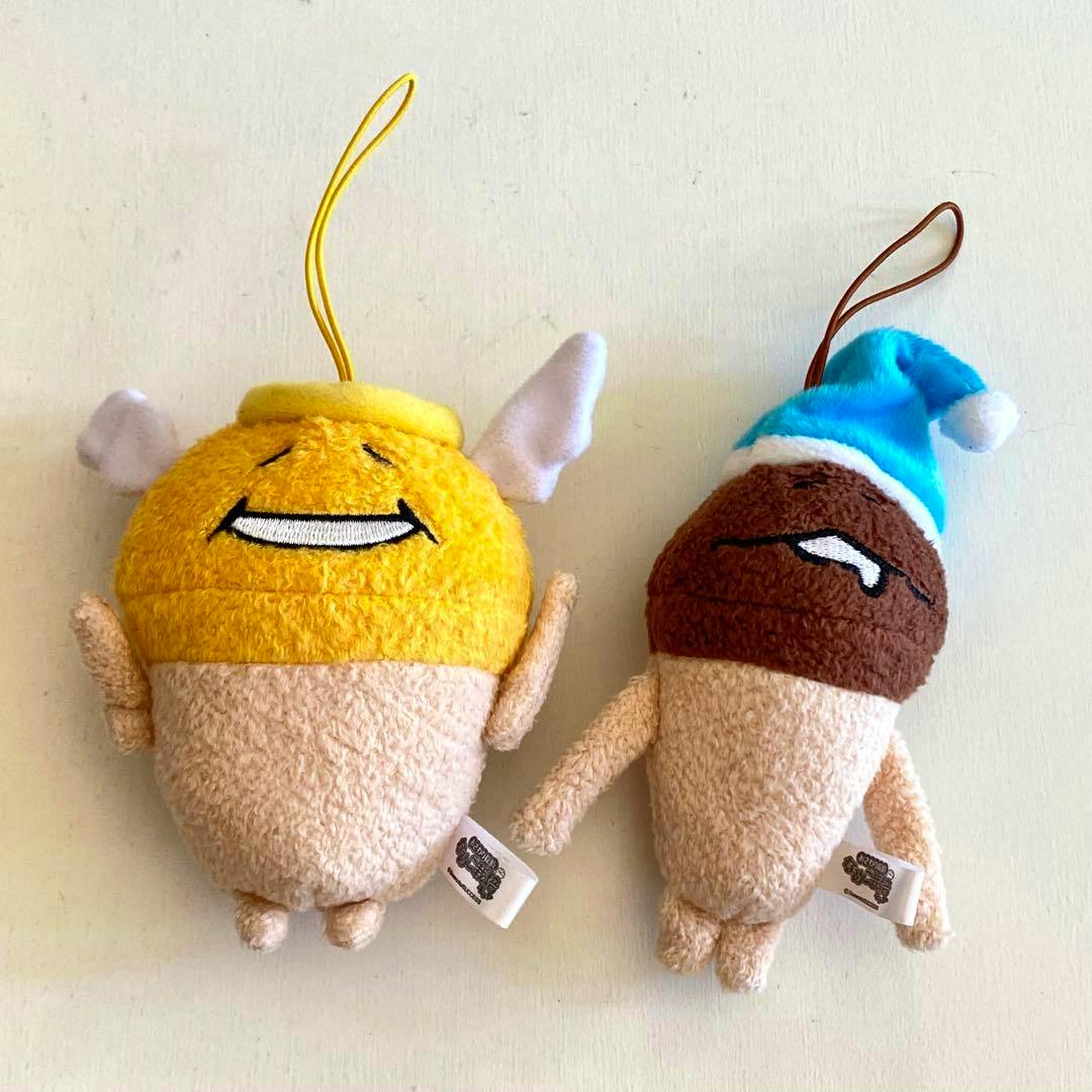 Front view of two Nameko cultivation kit plush mascots: a yellow one with small wings and halo-like loop and a brown one wearing a blue hat, both with hanging loops.