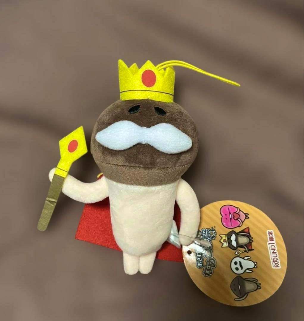 Front view of the "王様なめこ" plush toy showing a yellow crown, white mustache, red cape, scepter, and an attached product tag.