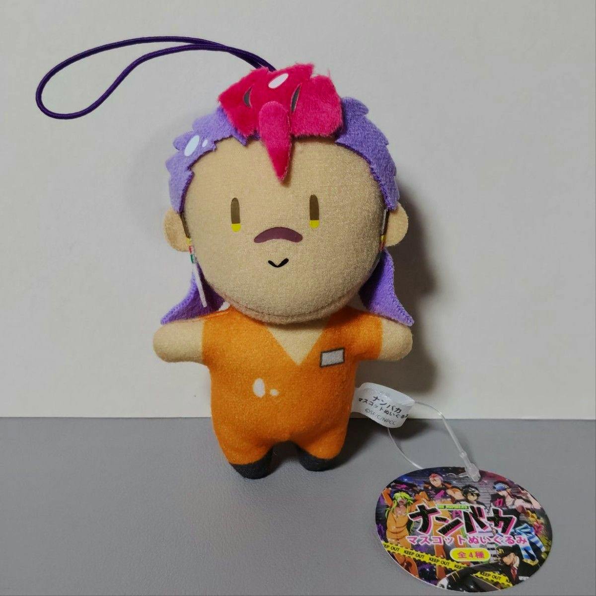 Front view of the タグ付き ナンバカ ロック マスコットぬいぐるみ 双又翔 plush toy wearing an orange outfit with purple and pink hair and an attached round tag.