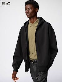 Model wearing the ポケッタブルUVカットパーカ hooded zip-up parka in black over a tan shirt and dark jeans.