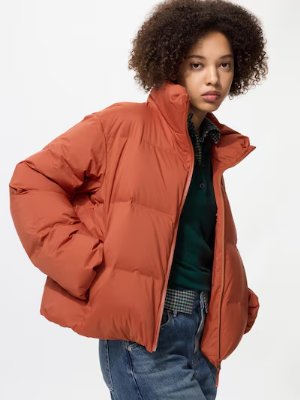 A woman modeling an orange ultralight down women's jacket against a white background.