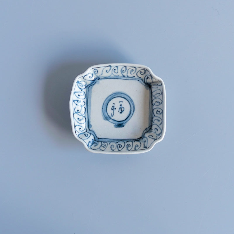 Top-down photo of the NAOKO UENO FUKUMOJI square ceramic plate with blue hand-painted decoration on a light background.