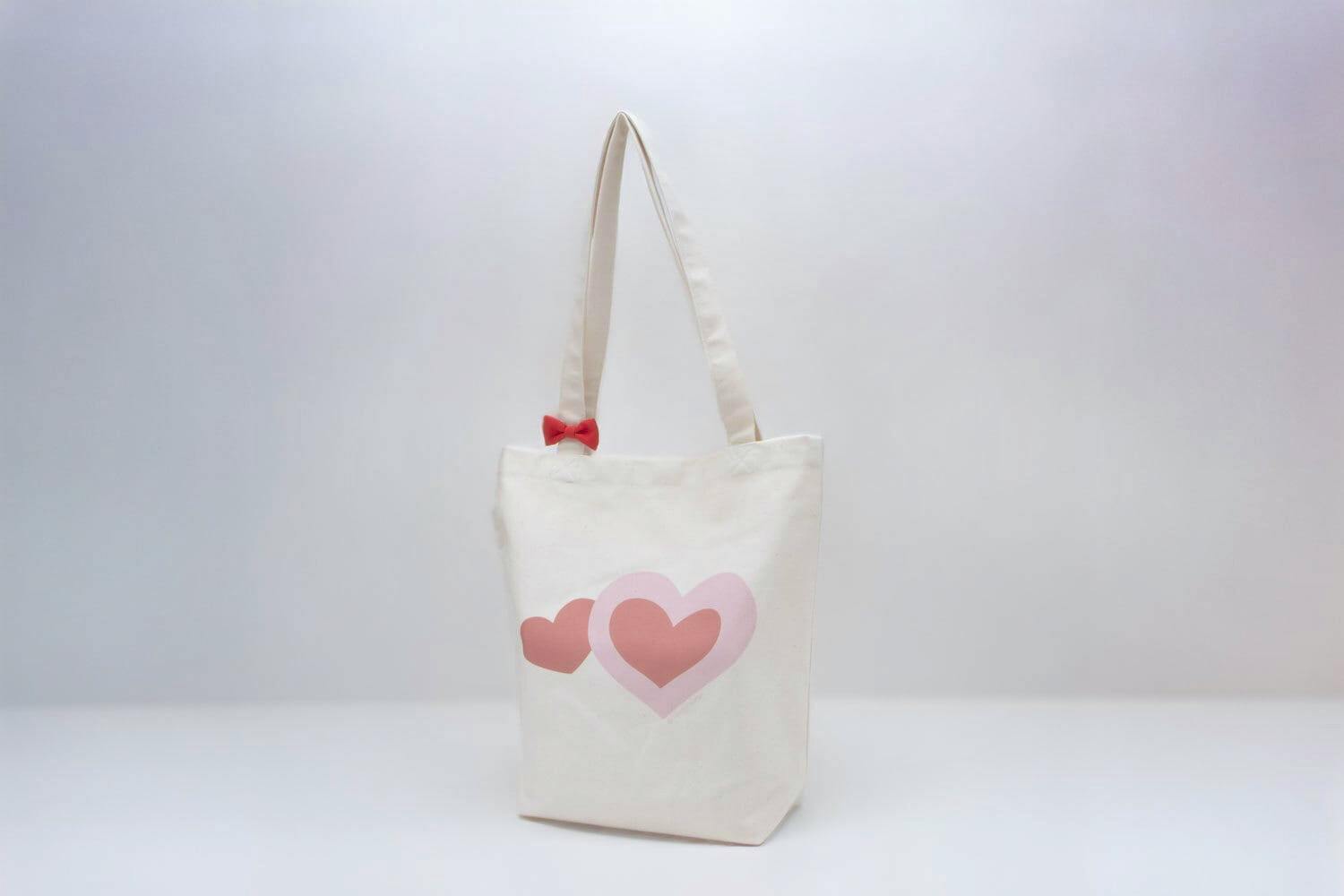 A white canvas tote bag with two pink hearts printed on the front and a small red bow on one strap.