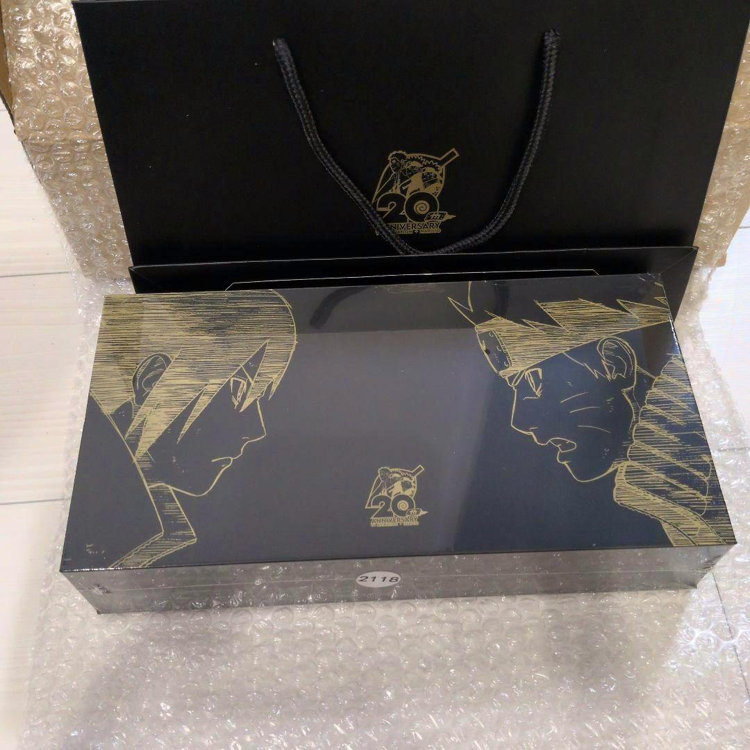 Sealed black Naruto 20th anniversary special collection box with gold line-art of two faces and a small 20th anniversary logo, shown on bubble wrap with a black gift bag behind.