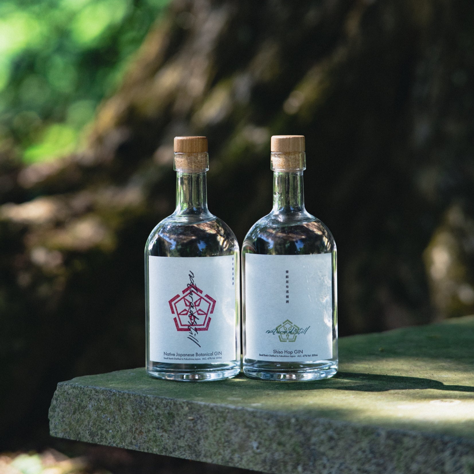 Two clear glass bottles of naturadistill Shiso Hop GIN with cork stoppers and white labels placed on a stone surface outdoors.