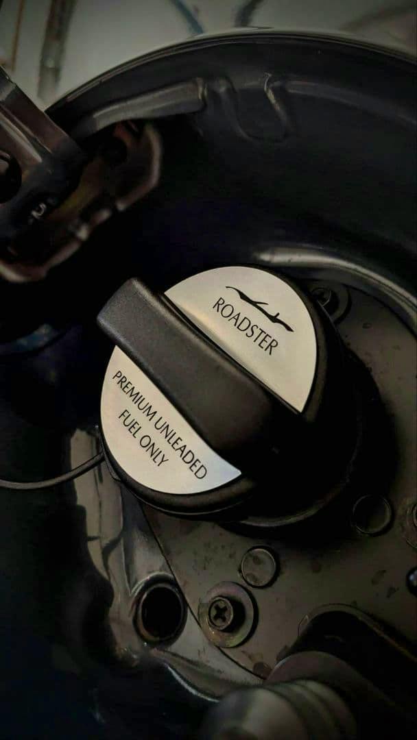 Installed ND Roadster fuel cap decoration in an aluminum hairline finish showing 'ROADSTER' and 'PREMIUM UNLEADED FUEL ONLY' inside a car fuel filler.