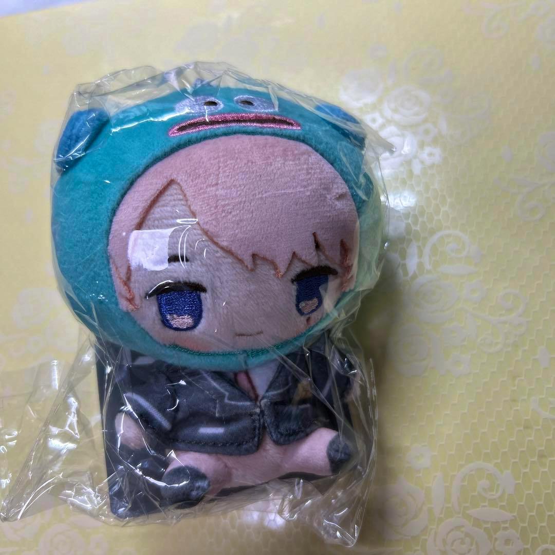 A clear-plastic-wrapped Mizunase Hangyodon plush keychain wearing a teal hood with embroidered face and blue eyes on a yellow patterned background.