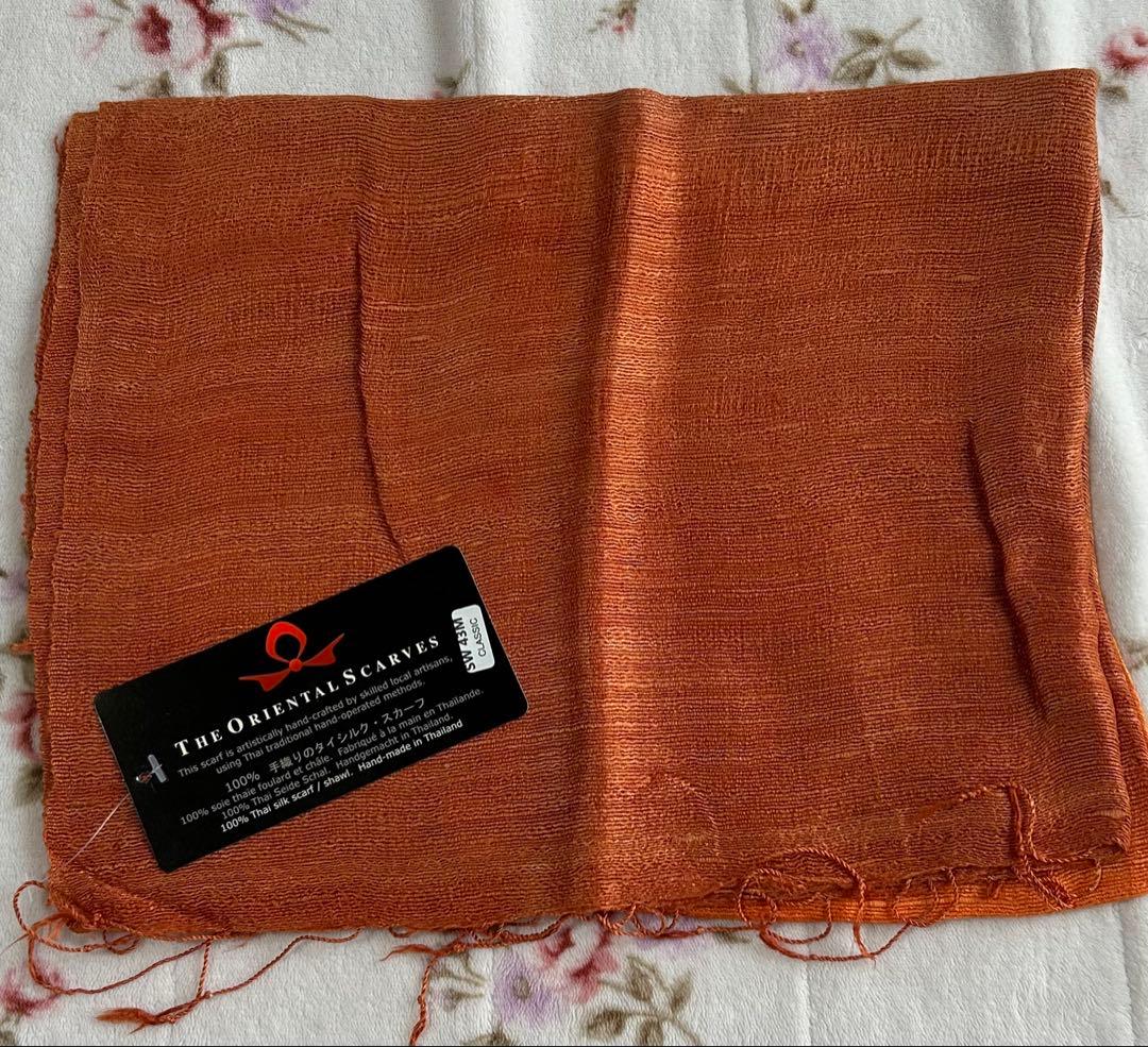 Orange handwoven Thai silk scarf laid flat with a black product tag visible.