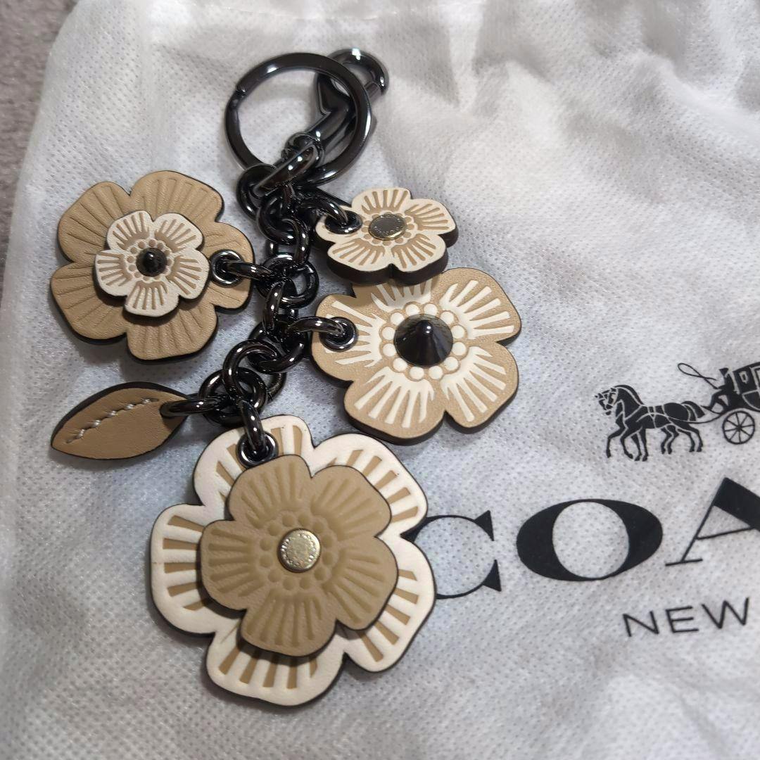Beige and white flower bag charm keychain with metal chain and ring displayed on a COACH dust bag.