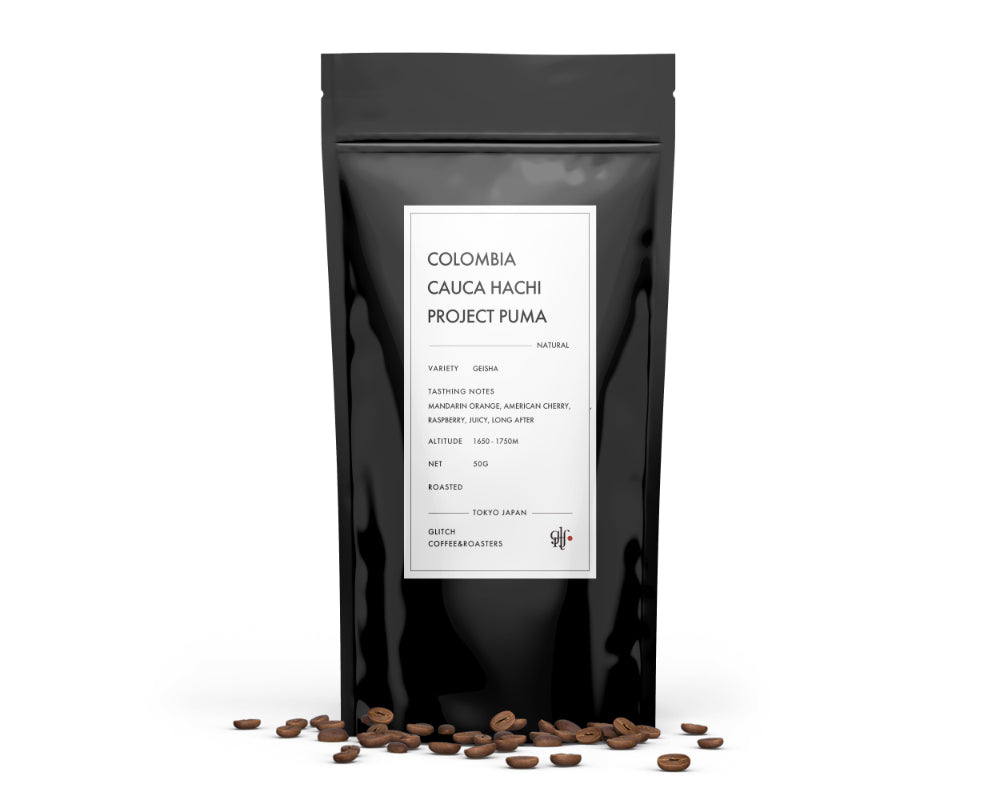 A black resealable coffee bag labeled "Colombia Cauca Hachi Project Puma" standing with roasted coffee beans scattered in front.