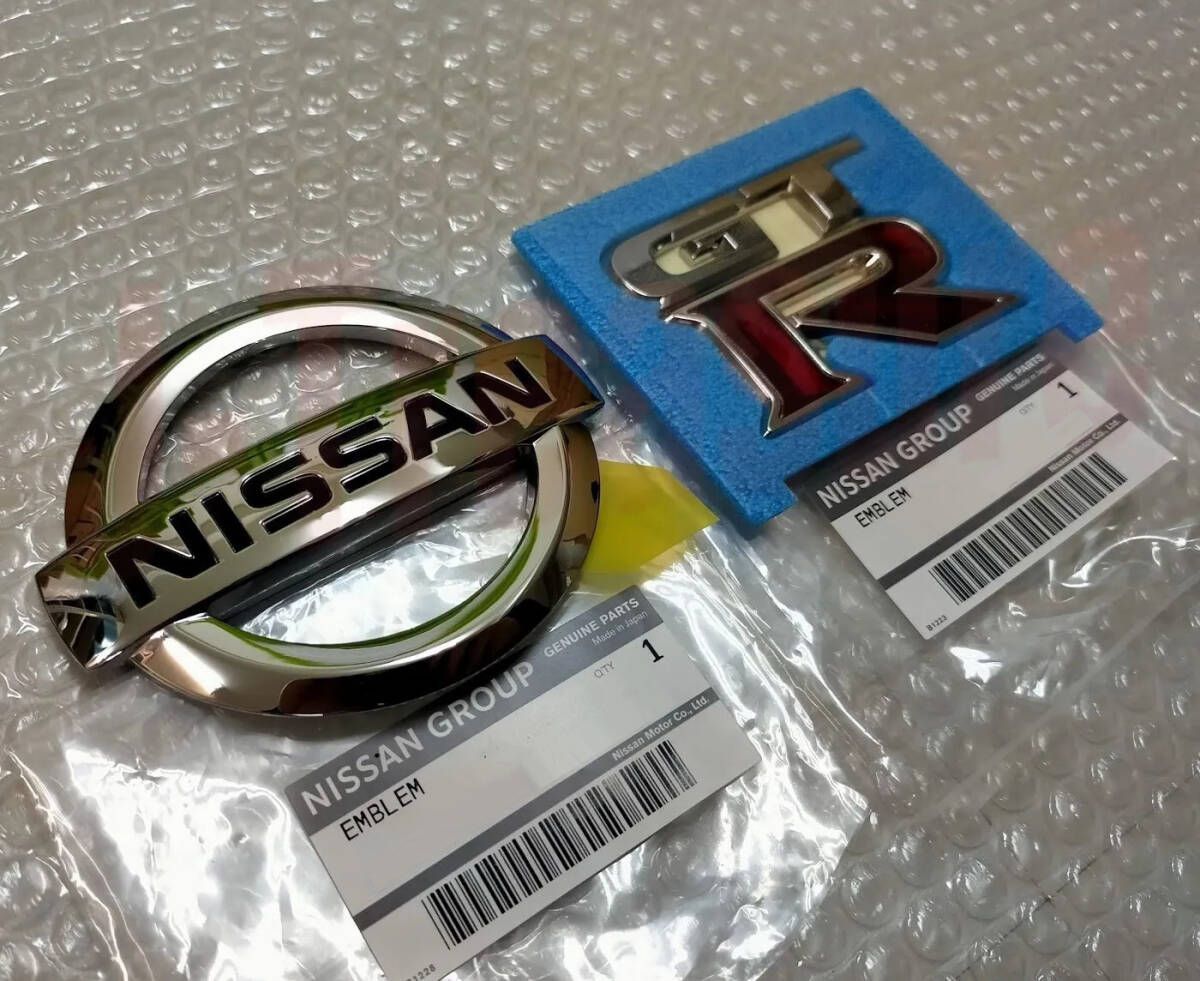 Two Nissan emblems (a round NISSAN badge and a red GT‑R badge) shown on bubble wrap with Nissan Group parts labels.