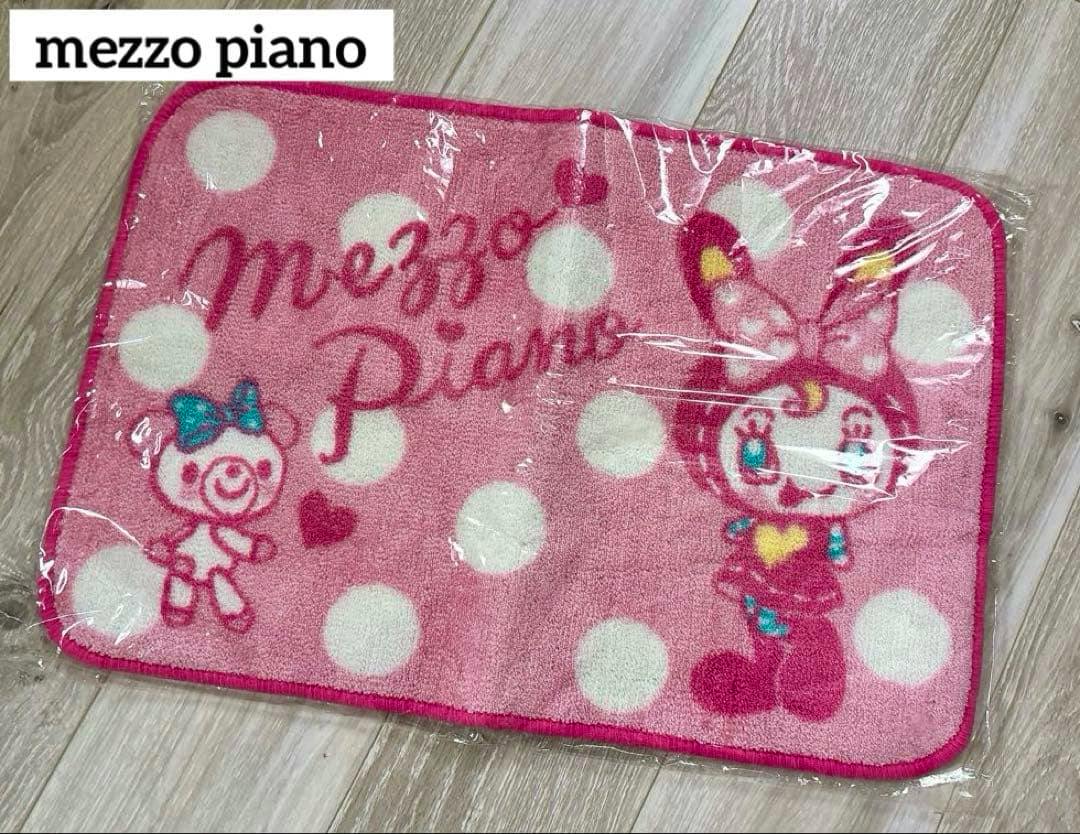 Pink polka-dot mezzo piano bath mat with character illustrations shown on a floor inside plastic wrapping.