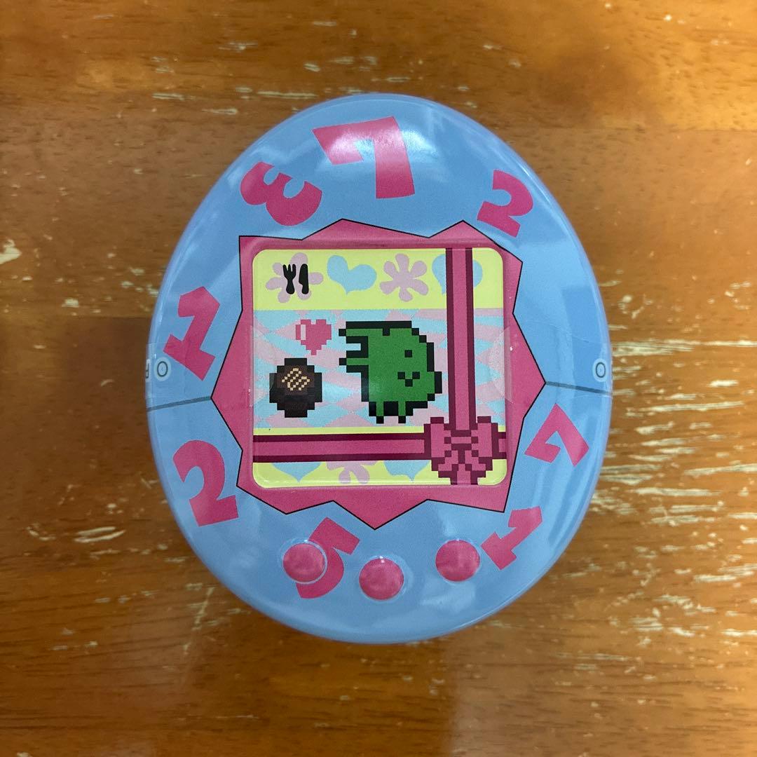 Light blue egg-shaped metal canister showing a pixelated Tamagotchi character and pink numbers on the lid.