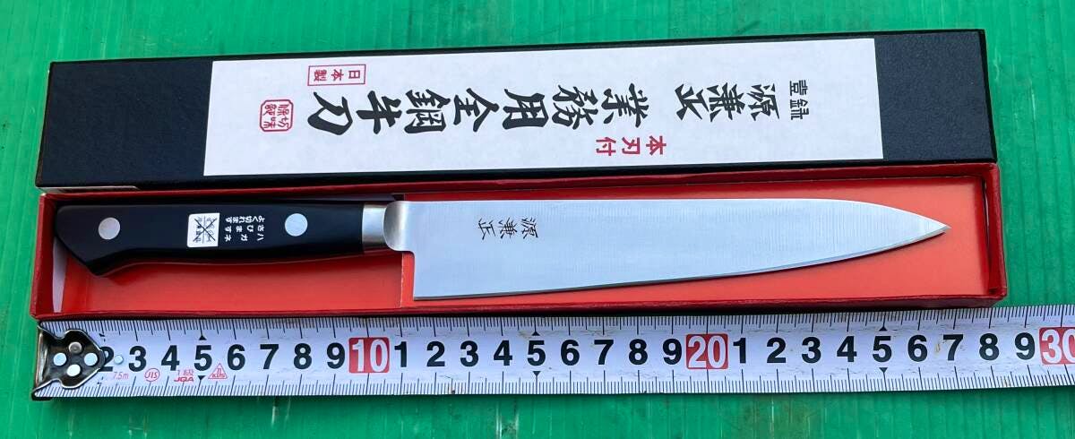 Chef knife with a black handle and exposed blade displayed in its open box with a measuring tape below.