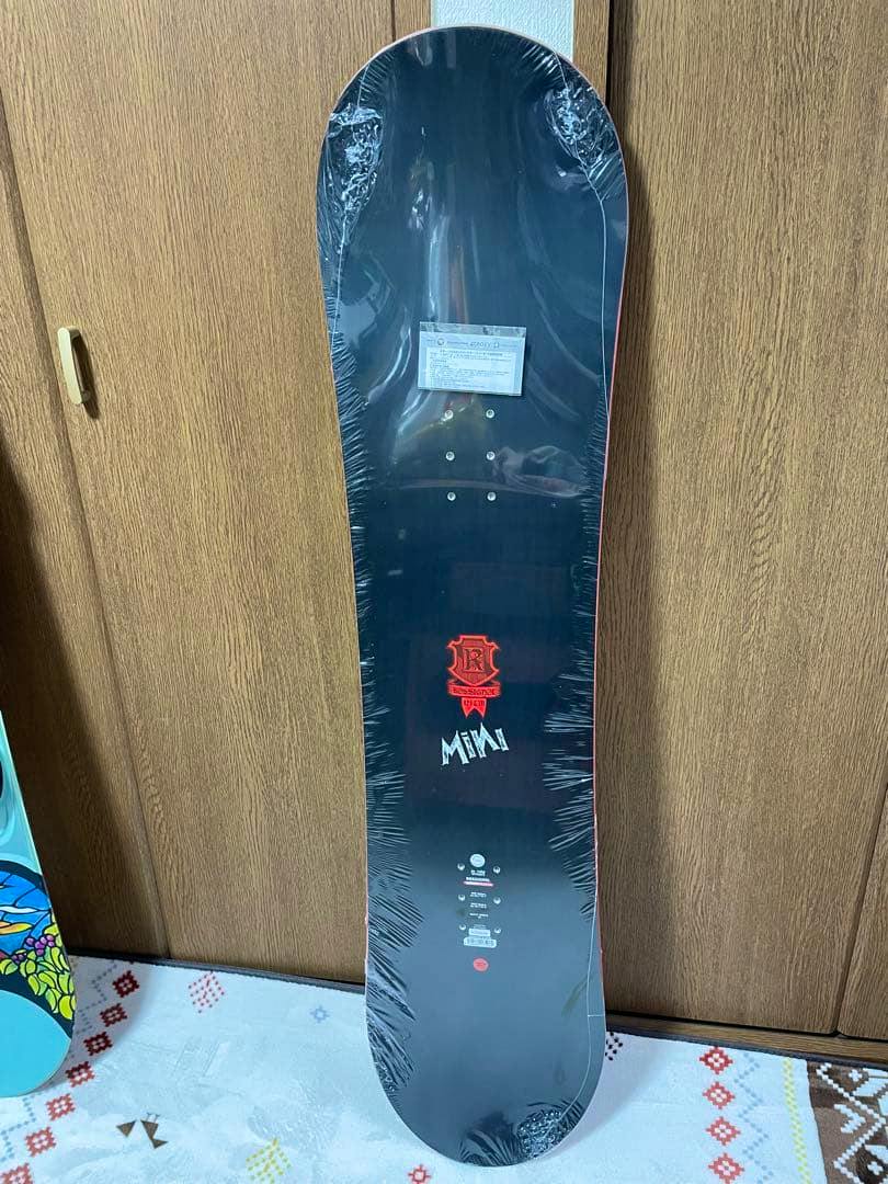 A wrapped snowboard from the listing '新品未使用 スノーボード 121cm' standing upright against a wooden door, showing the top sheet graphics and binding inserts.