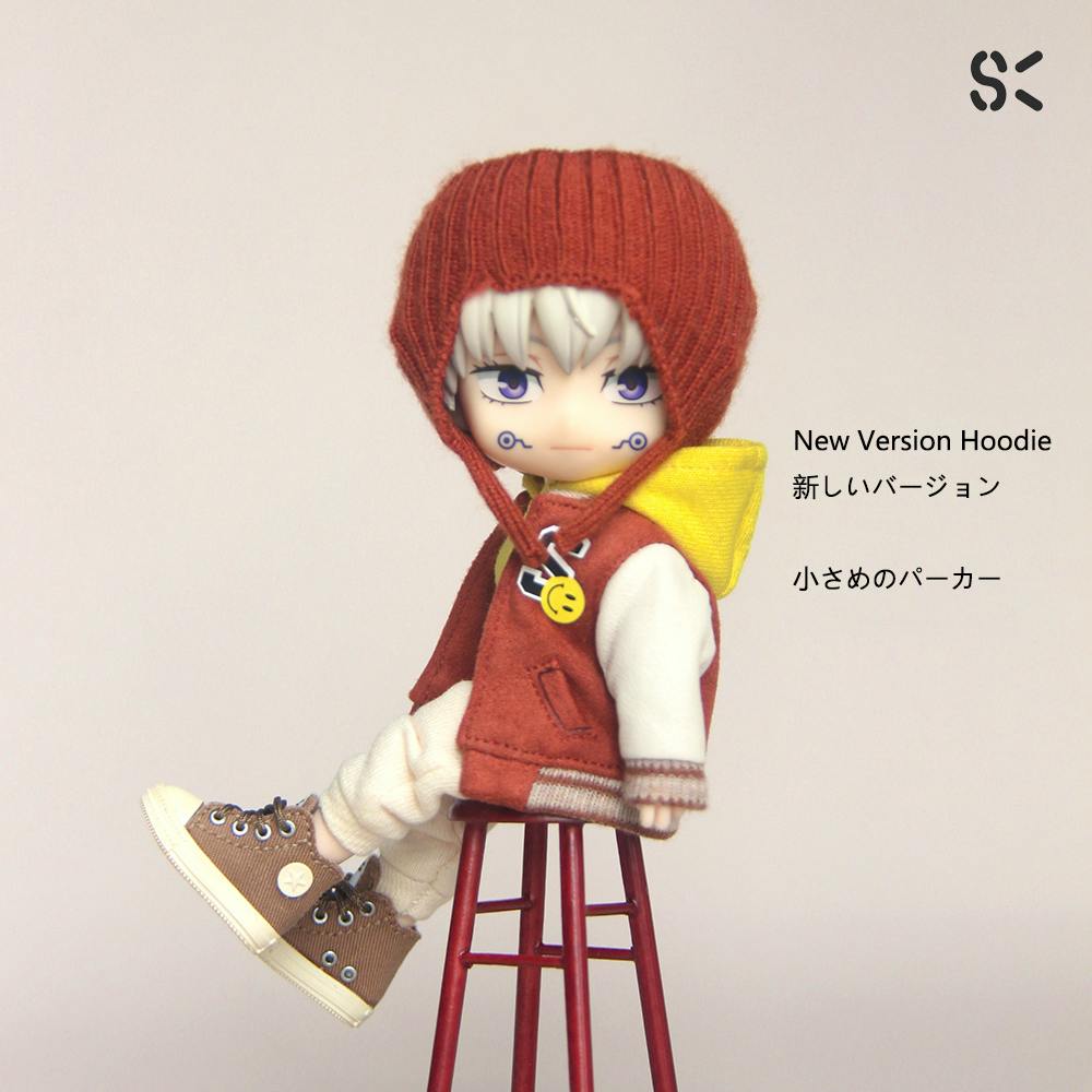 A doll wearing the hoodie and sweatpants set with a rust-colored jacket and yellow hood, cream pants, and brown sneakers seated on a red stool.