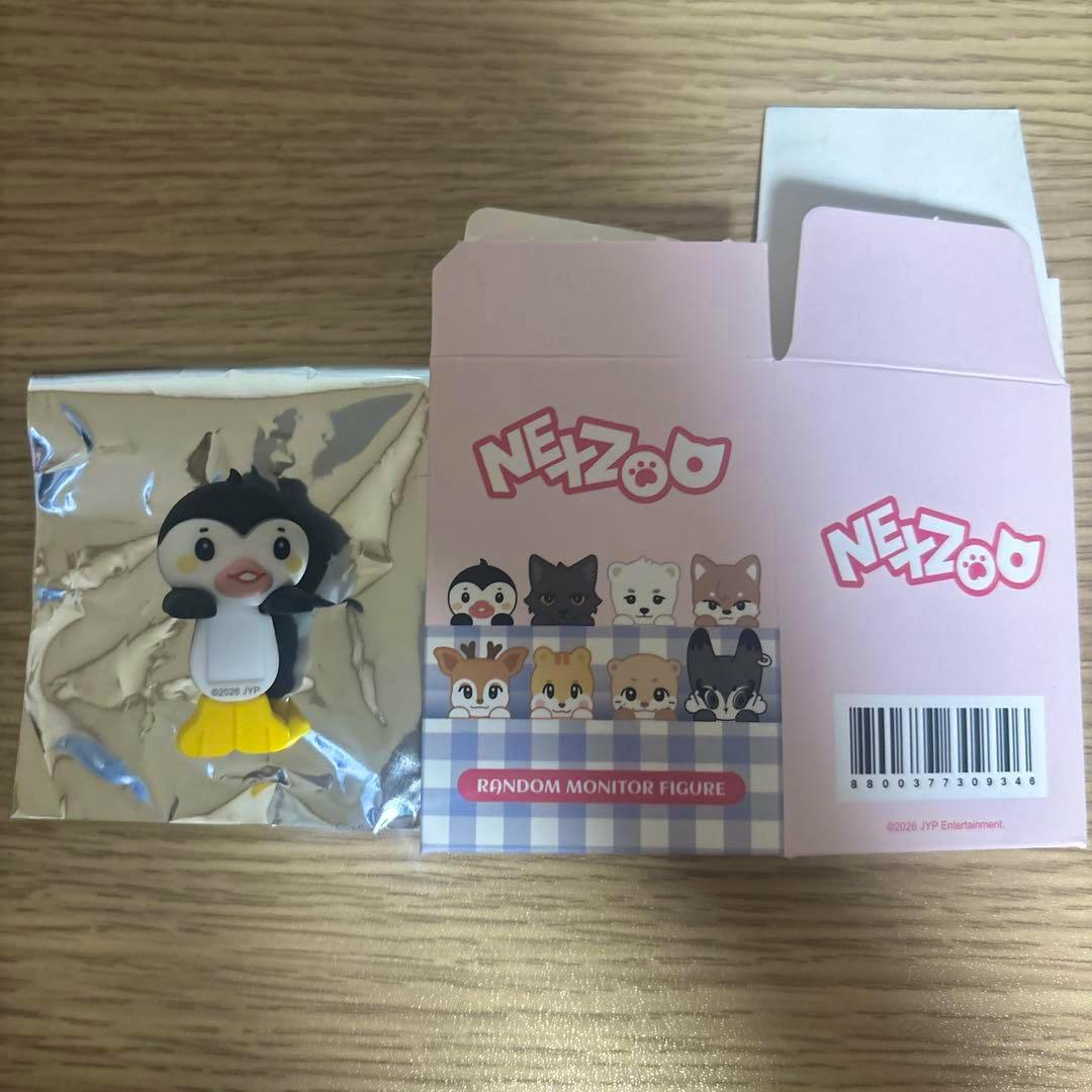 A small penguin monitor figure in foil packaging next to an opened pink NEZOO product box on a wooden surface.