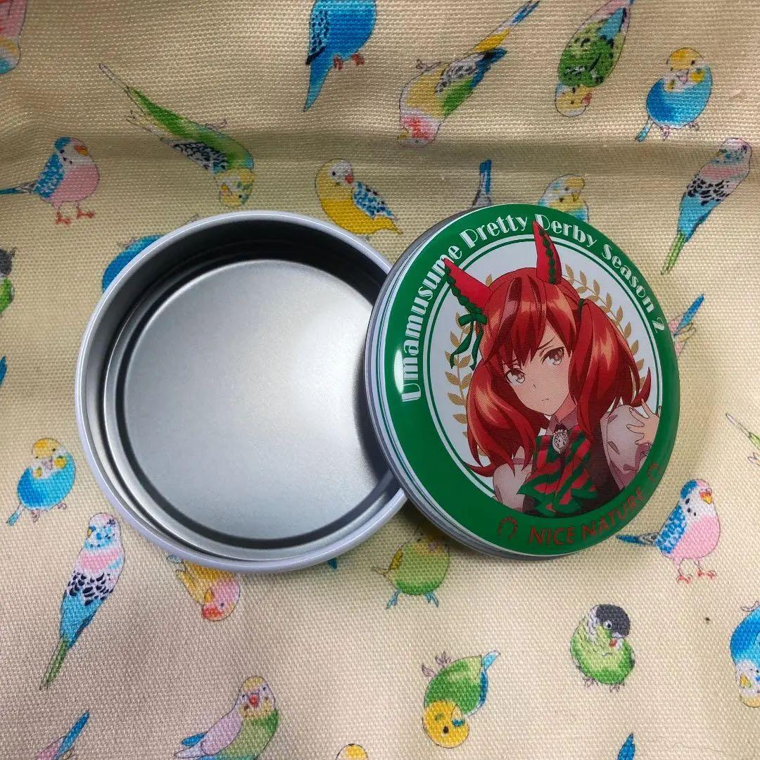 Open empty round metal can and its illustrated lid featuring ナイスネイチャ (Nice Nature) from ウマ娘 プリティーダービー, on patterned fabric.