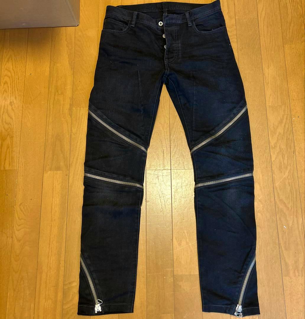 Front view of men's dark denim zipper jeans laid flat on a wooden floor, showing button fly and diagonal silver zippers.