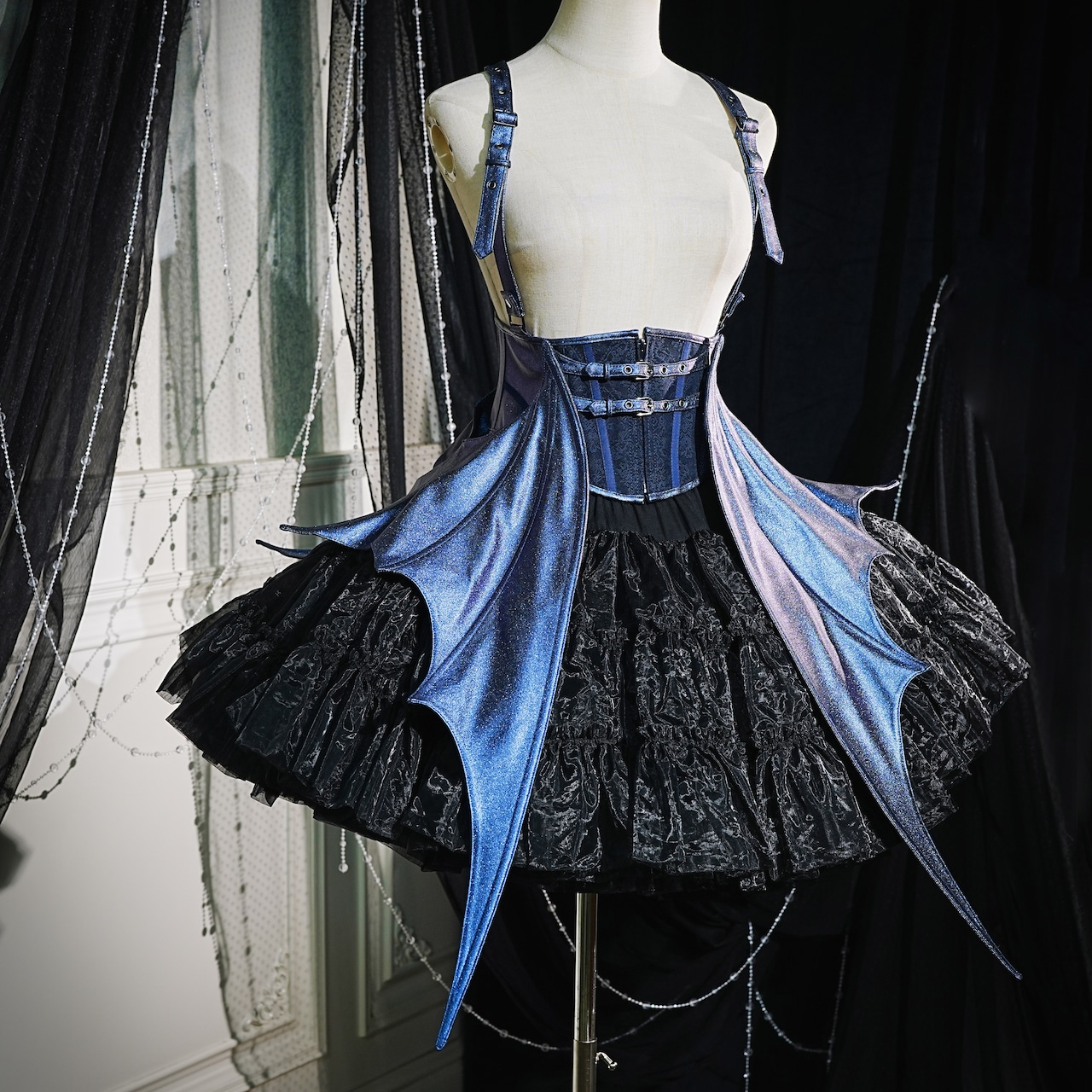 Blue harness corset with long wing-like side panels displayed on a dress form over a black ruffled skirt.