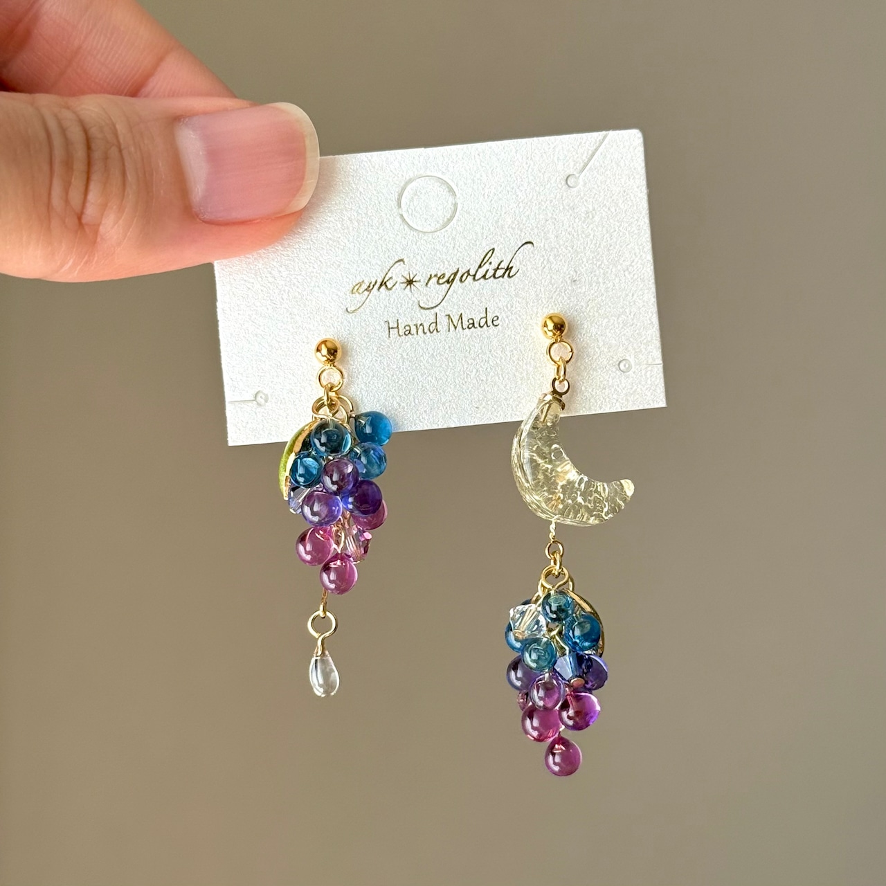 Pair of Night Sky Grapes earrings in blue and purple displayed on a white card held by a hand.